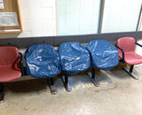 Four office chairs in a row, with three covered in blue protective plastic bags and one exposed red chair on the right, set in an indoor waiting area with tiled walls and windows.